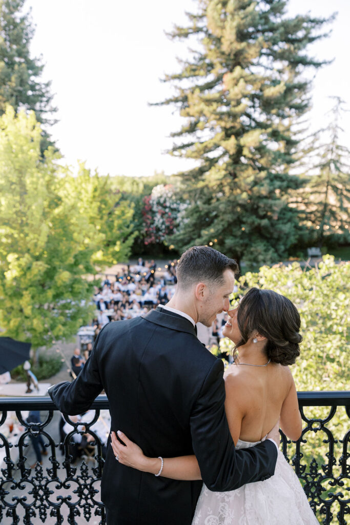 A couple in formal attire stands on a balcony, embracing and looking at each other, with a wedding reception and trees visible in the background.
