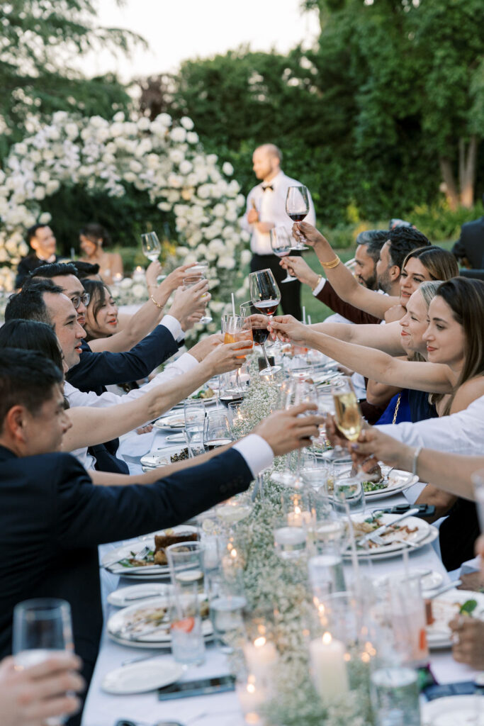 A group of people seated outdoors at a long dining table raise their glasses for a toast, with a flower arch and greenery visible in the background.
