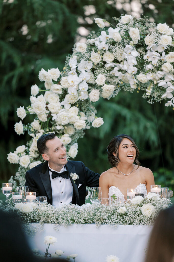 A bride and groom sit at a decorated table, smiling and looking to the side, with a white floral arch and greenery in the background.