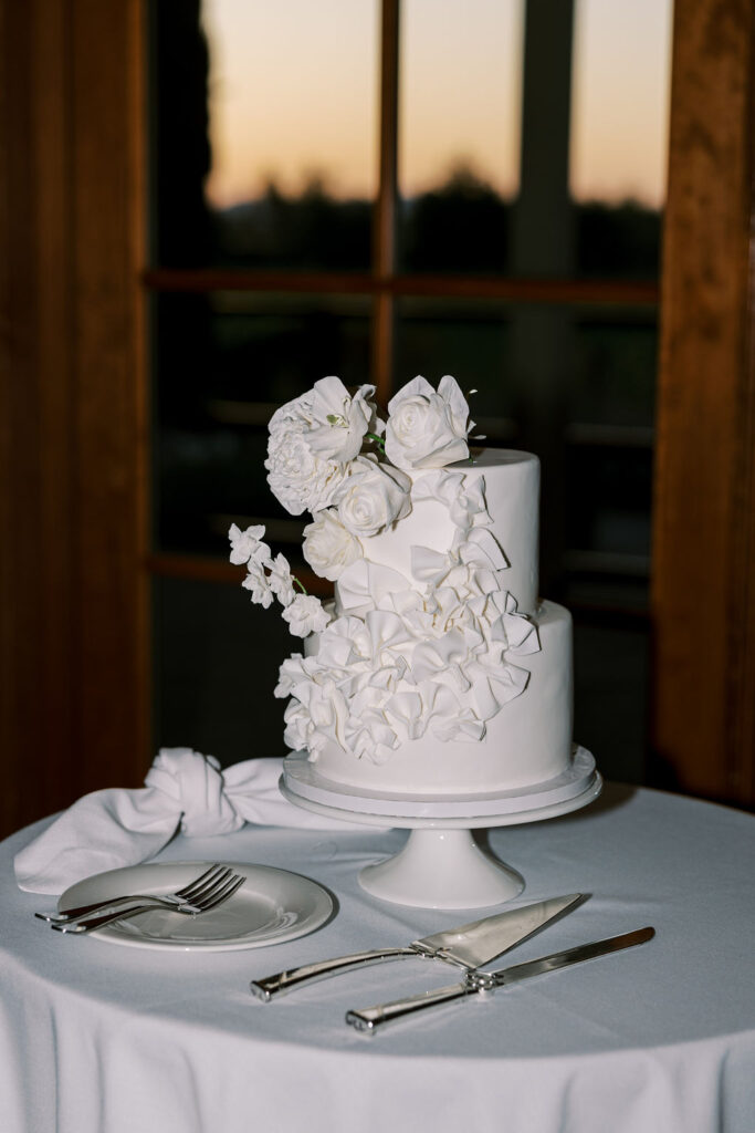 A two-tier white cake decorated with white flowers sits on a cake stand on a round table with a napkin, fork, knife, and cake server.