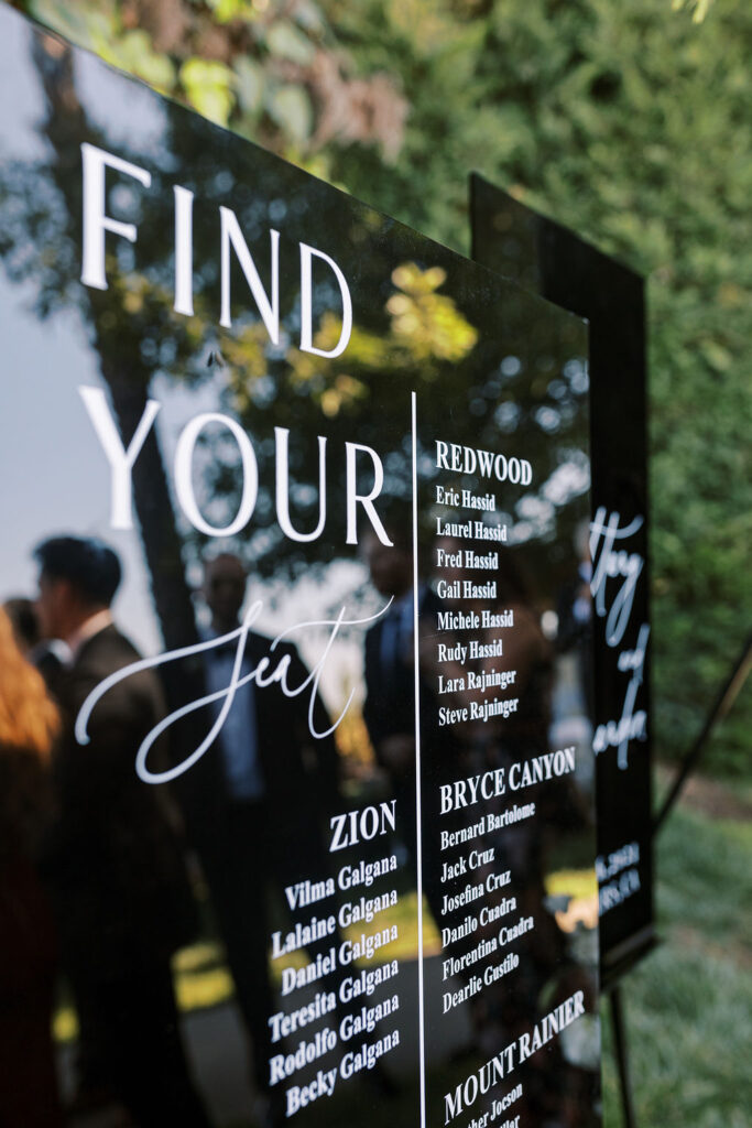 A glossy black seating chart display with white text lists guest names under categories like "Redwood" and "Zion," with greenery and people blurred in the background.