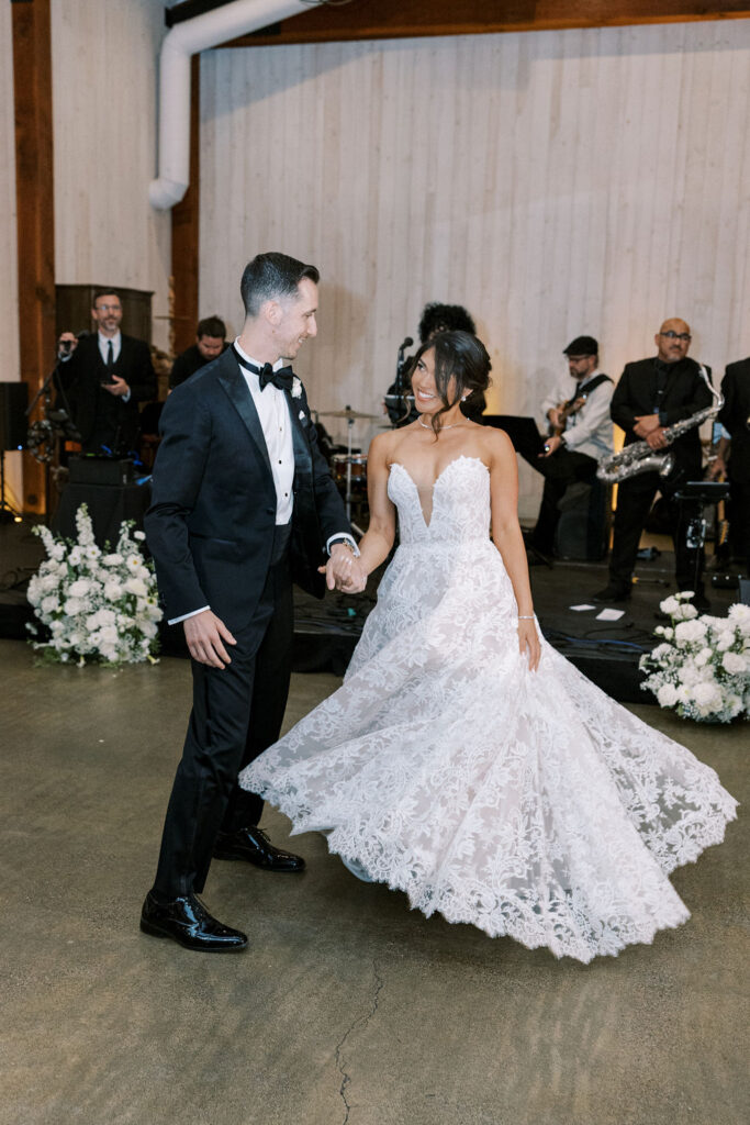 A bride and groom dance together at their wedding reception, with a live band performing in the background and floral arrangements on the floor.