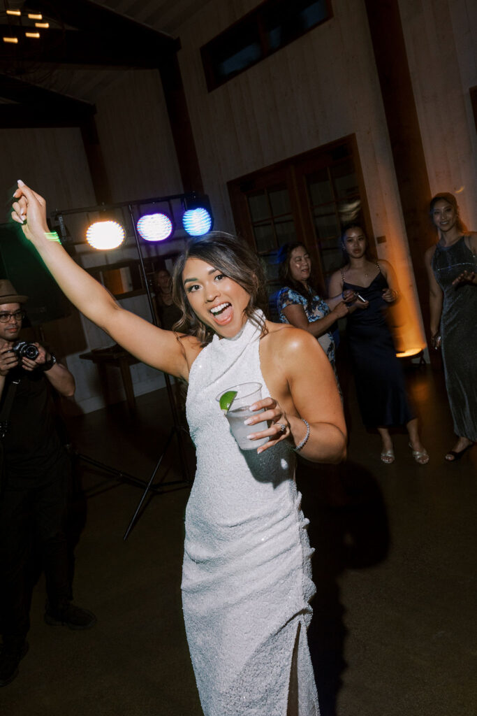 A woman in a white dress holds a drink and raises her arm while smiling at a party, with people and colorful lights in the background.