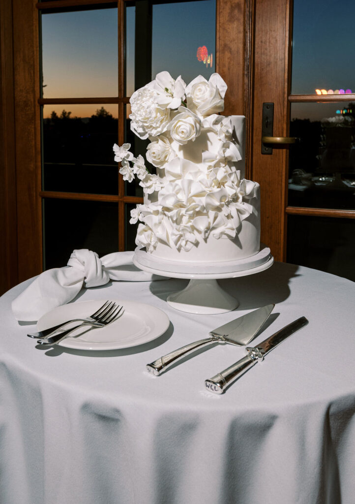 A white two-tiered cake decorated with white flowers sits on a round table set with a napkin, plate, fork, knife, and cake server in front of glass doors at sunset.