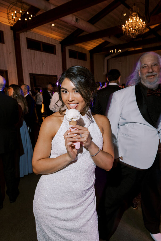 A woman in a white dress holds and licks an ice cream cone at an indoor event, with guests and chandeliers visible in the background.