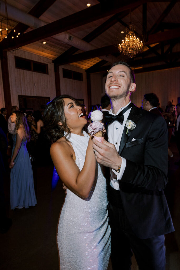 A woman in a white dress and a man in a black tuxedo smile and laugh together while holding an ice cream cone at an indoor event.