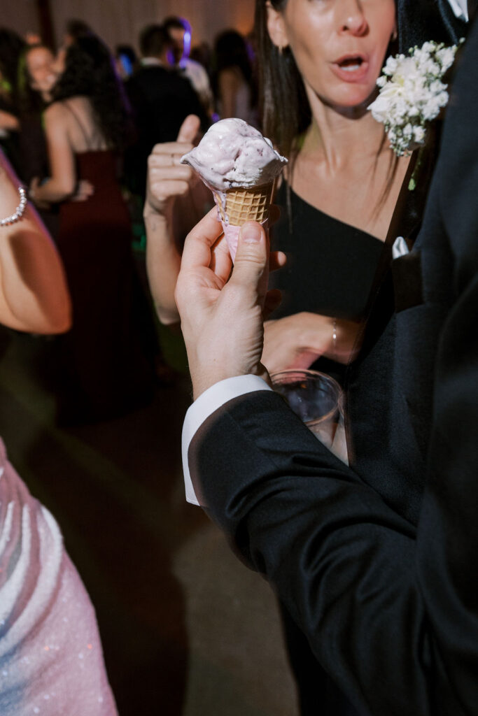 A person in formal attire holds an ice cream cone at an event, surrounded by other guests engaged in conversation.