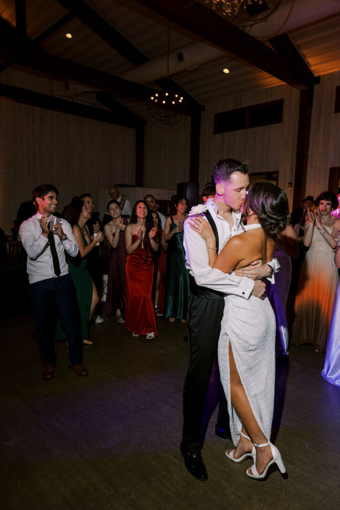 A couple shares a kiss while dancing at an indoor event, surrounded by guests in formal attire who are clapping and watching.