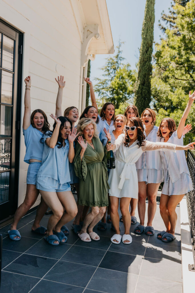 A group of women, some in matching light blue pajamas and slippers, stand on a patio cheering and smiling around a woman in a white robe and a woman in a green robe.