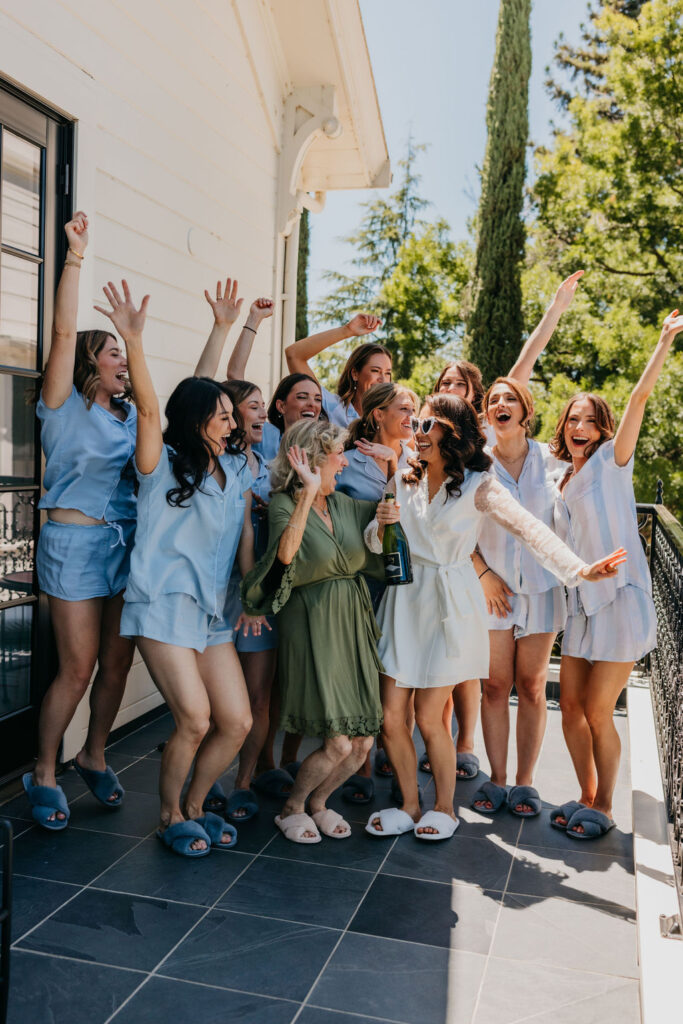 A group of women, some in matching blue pajamas and slippers, celebrate on a patio with one woman in white holding a champagne bottle.