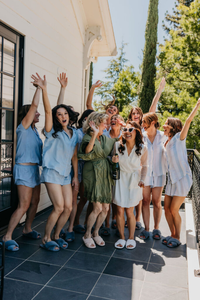 A group of women in sleepwear and slippers stand on a patio, smiling and celebrating with raised hands and a bottle of champagne.