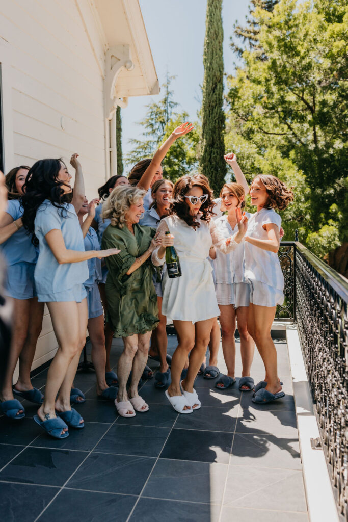 A group of women in matching outfits celebrate outdoors on a balcony, with one woman holding a bottle of champagne and wearing sunglasses.