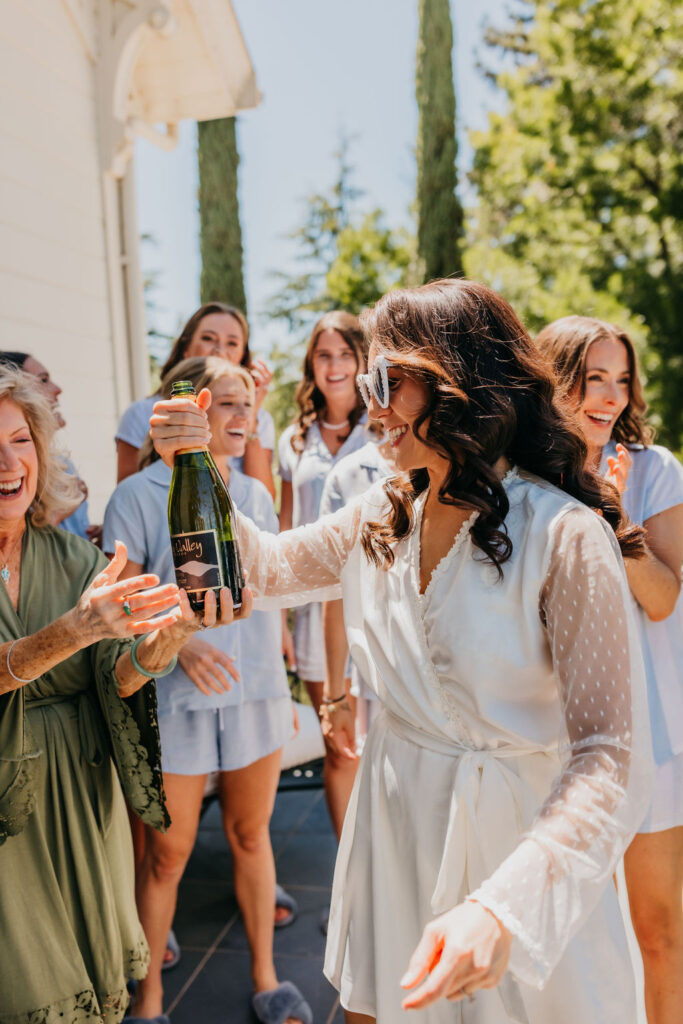A woman in a white robe and sunglasses holds up a bottle of bubbly, surrounded by smiling women outdoors on a sunny day.