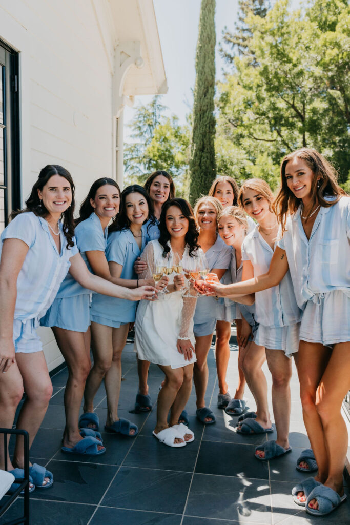 A group of women in matching blue pajamas and slippers stand on a patio, smiling and holding drinks, with greenery in the background.