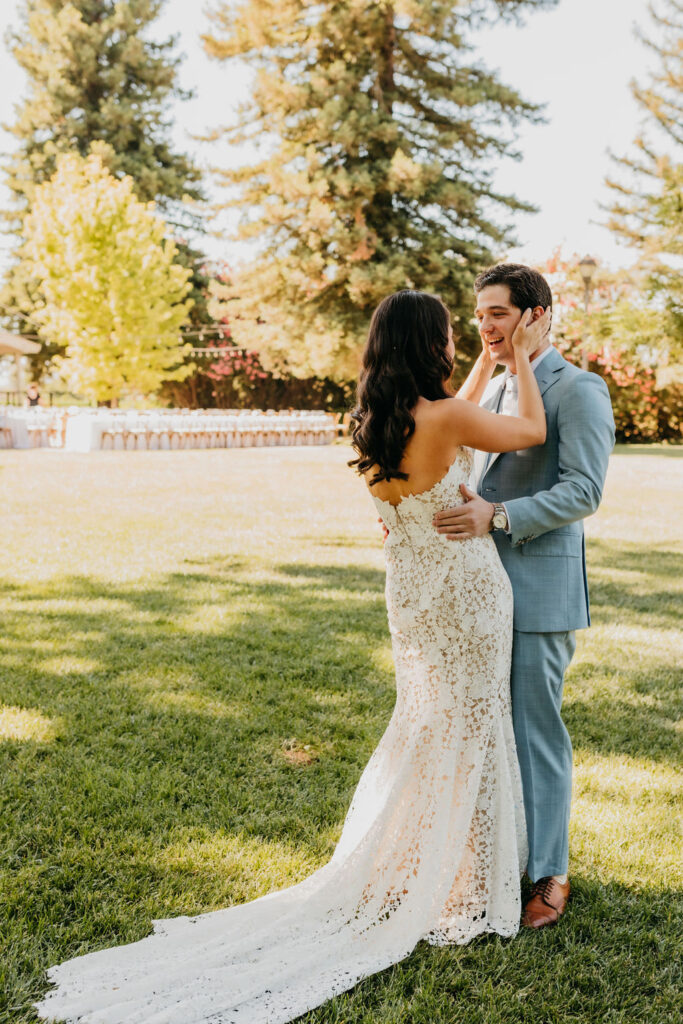 A bride in a lace dress and a groom in a light blue suit embrace and smile at each other outdoors on a sunny day, with trees and grass in the background.