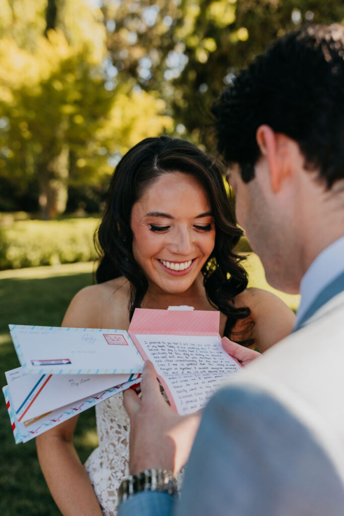 A woman in a white dress smiles while a man in a suit reads a handwritten letter to her outdoors, holding several colorful envelopes.