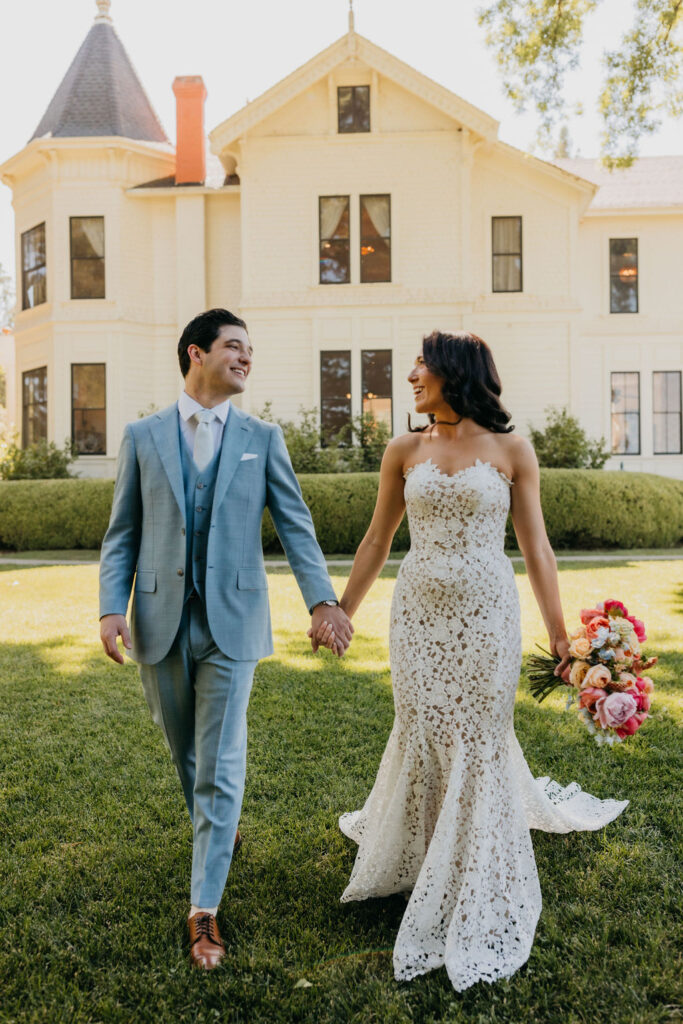A bride in a white lace dress and a groom in a light blue suit walk hand in hand on grass in front of a large, light-colored house.