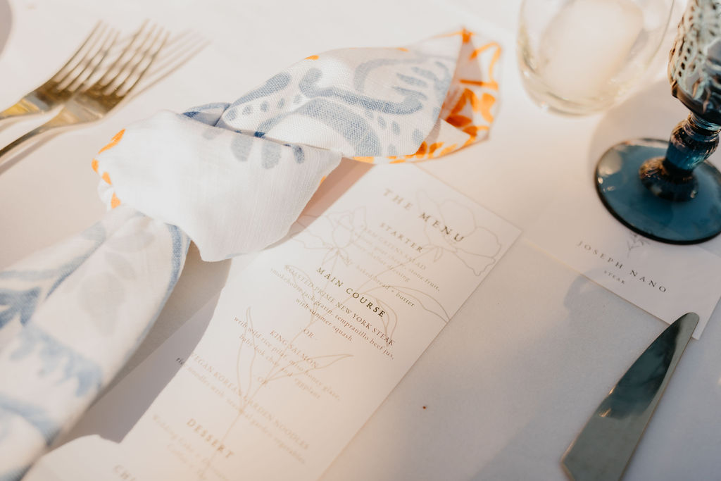 A close-up of a table setting with a printed menu, folded napkin, cutlery, a drink, and a place card on a white tablecloth.