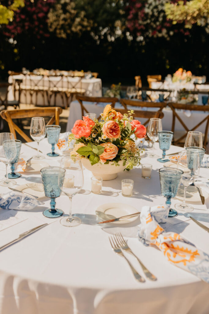 A round table set for an outdoor event features blue glasses, white plates, silver cutlery, and a floral centerpiece with orange and yellow flowers.