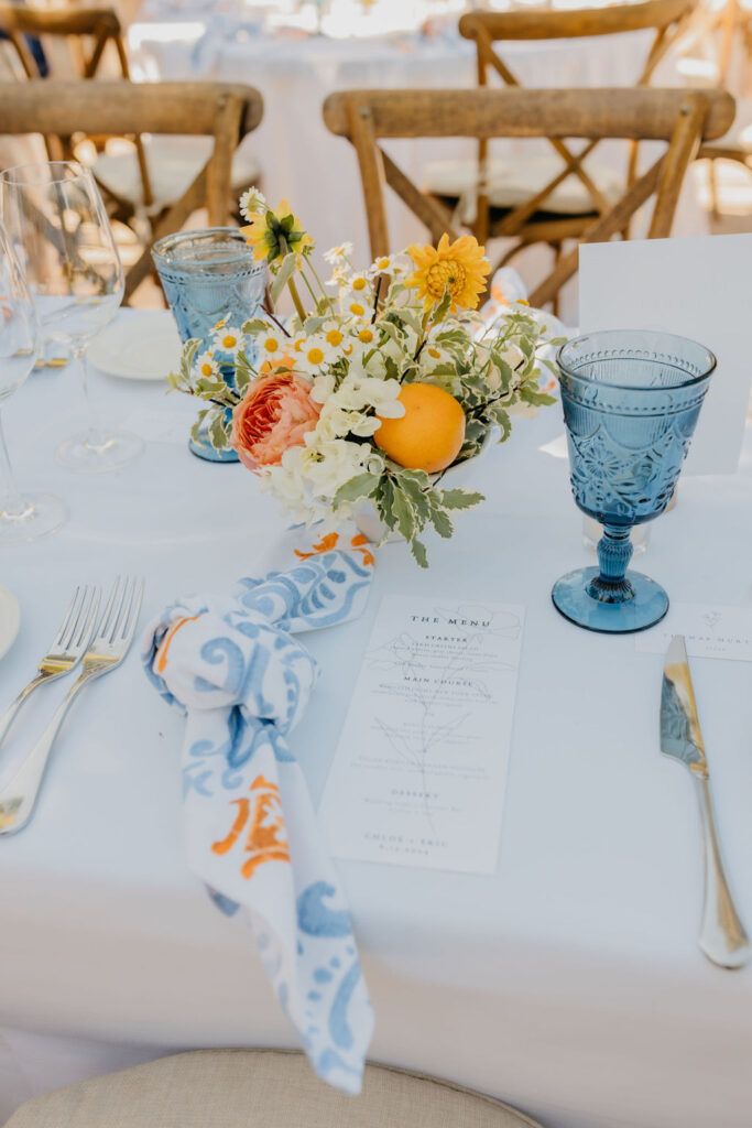 A table setting with blue glassware, gold cutlery, a floral centerpiece with citrus fruits, a printed menu, and a cloth napkin with blue and orange patterns.