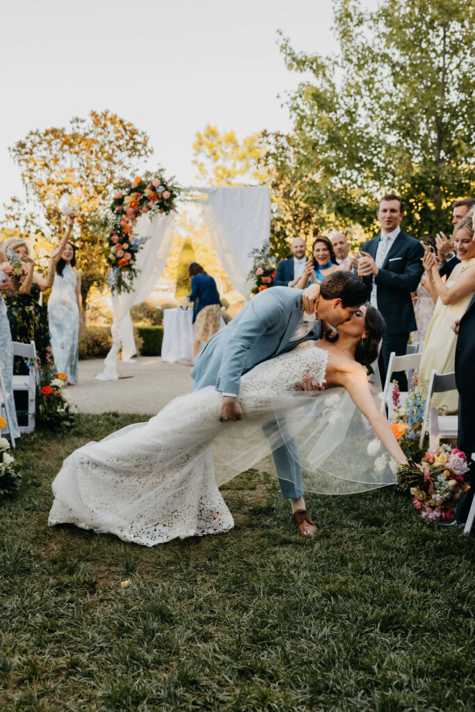 A groom in a blue suit dips and kisses the bride in a white lace gown during an outdoor wedding ceremony, with guests applauding and floral decorations in the background.