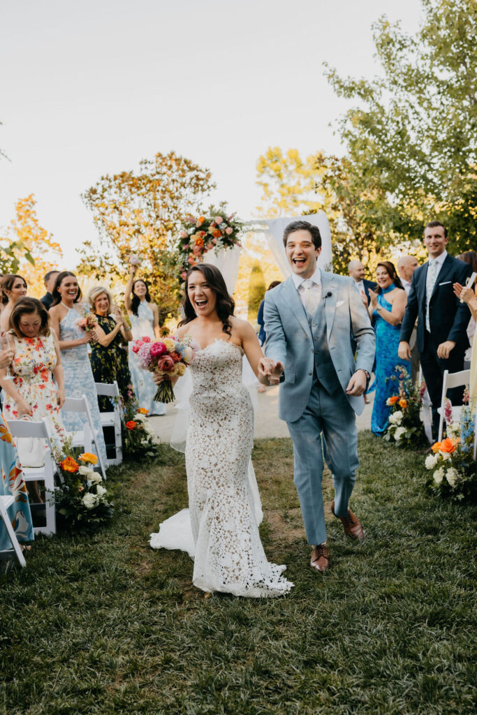 A newlywed couple walks down an outdoor aisle, smiling and holding hands, surrounded by cheering guests and floral decorations.