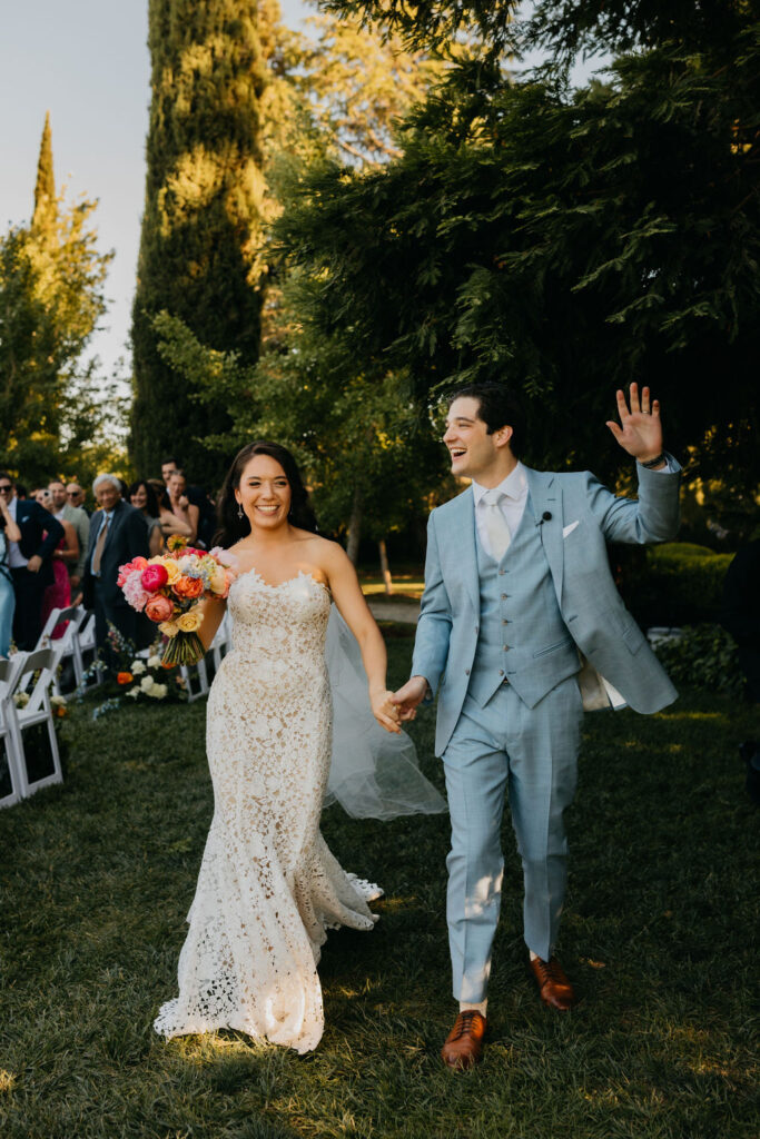 A bride in a white lace gown and a groom in a light blue suit walk hand in hand outdoors, smiling and waving, surrounded by wedding guests.
