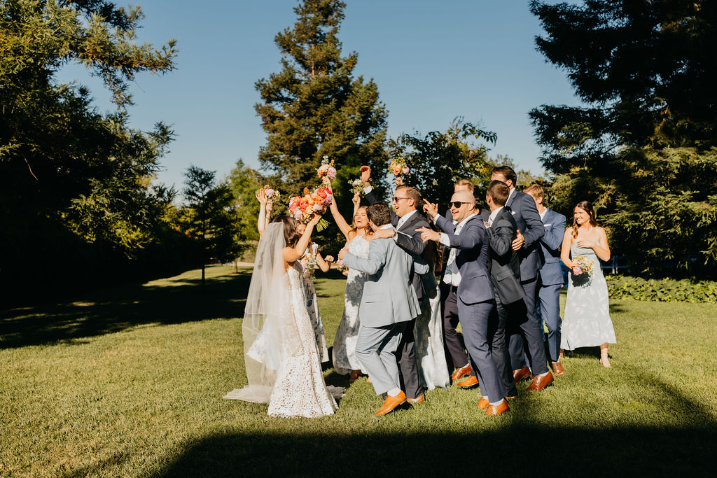 A wedding party in formal attire stands on grass, celebrating with raised arms and flower bouquets on a sunny day.