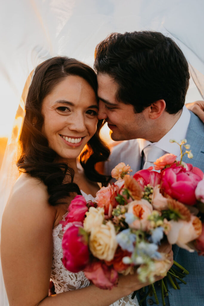 A bride smiles while holding a colorful bouquet as the groom leans in close, both under a sheer veil in natural light.