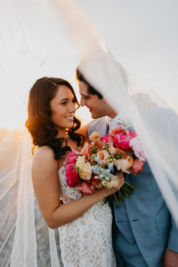 A bride in a lace dress and groom in a light blue suit stand close together, holding a colorful bouquet under a flowing veil at sunset.