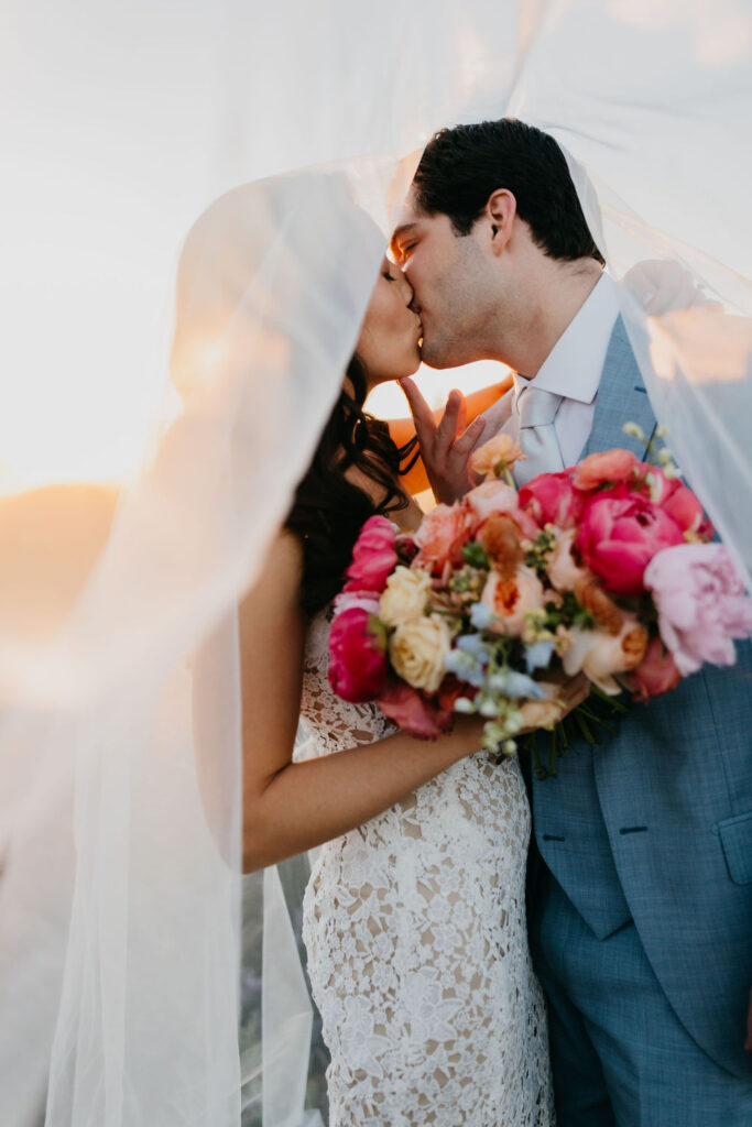 A bride and groom kiss under a sheer veil; the bride holds a colorful bouquet of flowers, and the groom wears a light blue suit.