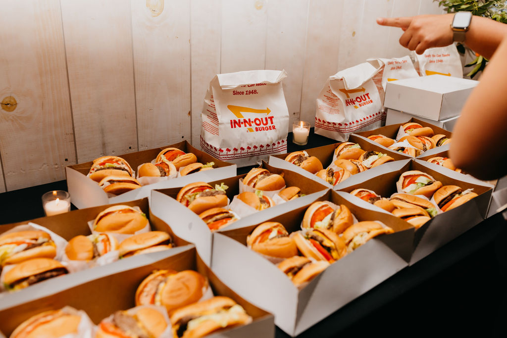 Multiple open boxes filled with In-N-Out burgers are arranged on a table; a hand points toward the food, and candles are visible in the background.