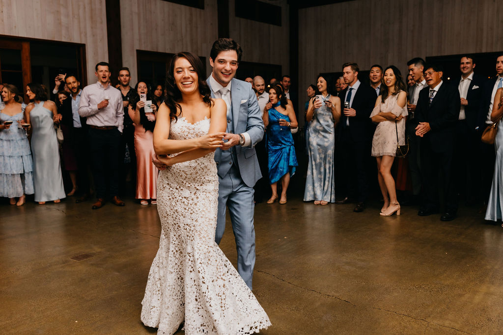 A newlywed couple, dressed in a white lace gown and light suit, dance together in the center of a room while a group of guests stand around watching and smiling.