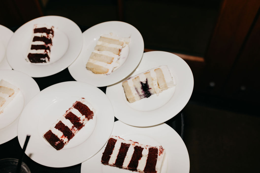 Several white plates with slices of layered cake, including red velvet and white cakes, arranged on a dark surface.