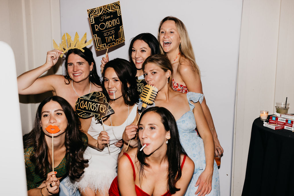 Six women pose together in a photo booth, holding props and signs, including "Have a Roaring Good Time" and "Gangster," with playful expressions and lollipops.