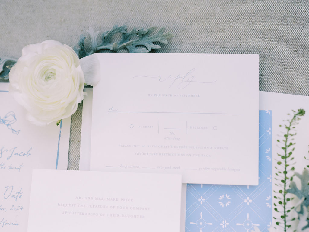 A flat lay of wedding stationery, including an RSVP card, an invitation, a blue patterned card, and a white ranunculus flower on a light fabric background.