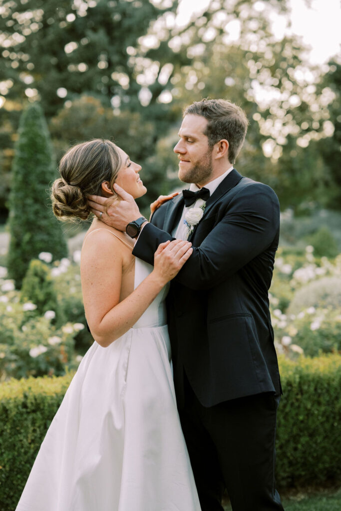 A bride and groom stand outside in a garden, gazing at each other and embracing, dressed in a white gown and black tuxedo.