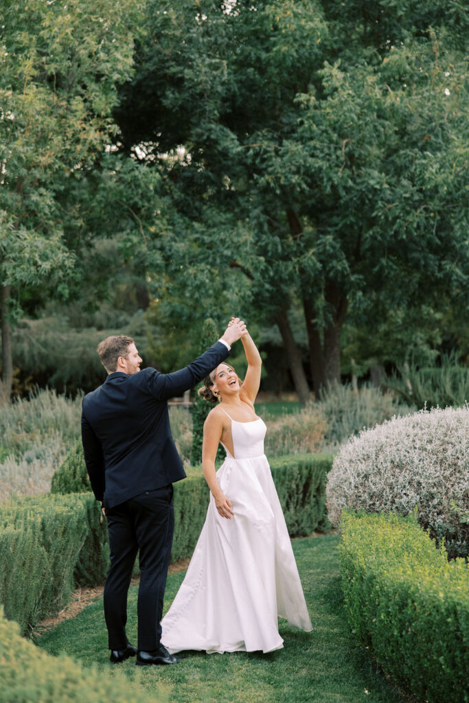 A bride in a white dress and a groom in a black suit dance together outdoors on a green lawn surrounded by bushes and trees.
