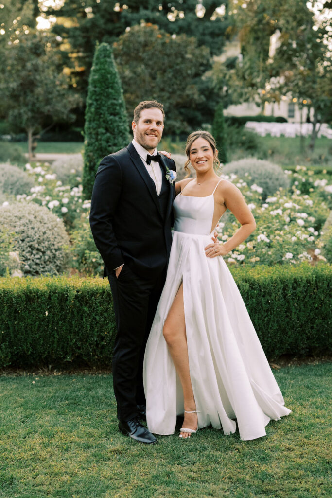 A man in a black tuxedo and a woman in a white gown pose together outdoors in a manicured garden with green hedges and flowering bushes.