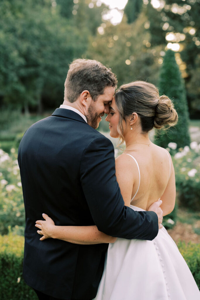 A bride and groom embrace outdoors, standing close with their foreheads touching, surrounded by greenery and white flowers.