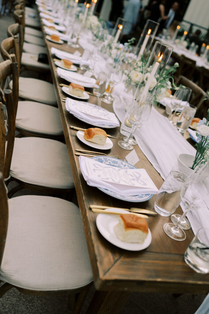 A long wooden table is set for a formal meal with plates, bread rolls, glasses, candles, and floral centerpieces.