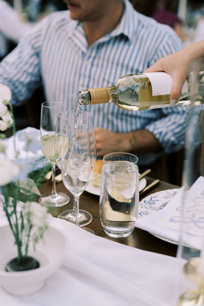A person pours white wine into a glass at a table set with water, wine glasses, white flowers, and a man in a striped shirt.