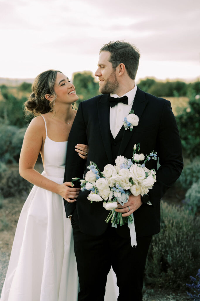 A bride and groom stand outdoors, the bride smiling at the groom who is holding a bouquet of white and blue flowers. Both are dressed in formal wedding attire.