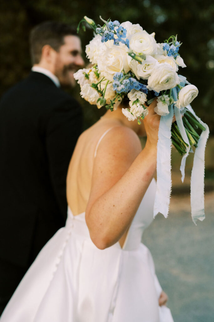 A bride in a white dress holds a bouquet of white and blue flowers, while a man in a black suit stands beside her, both outdoors.