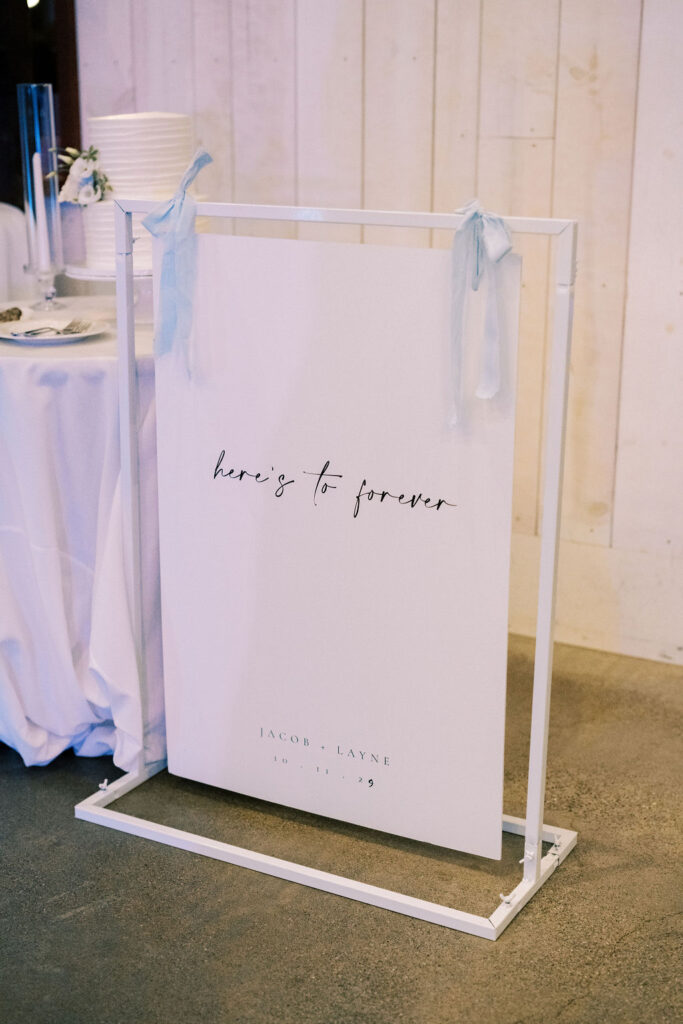 A white wedding sign with blue ribbon reads "here's to forever" and displays the names "Jacob + Layne" and the date "10 . 14 . 23" in a modern, minimal font.