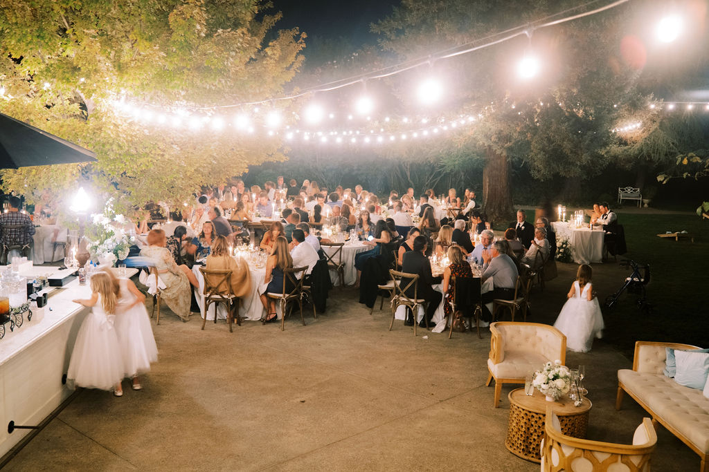 Outdoor evening wedding reception with guests seated at tables under string lights, children in white dresses, and decorated tables and lounge area on a patio surrounded by trees.