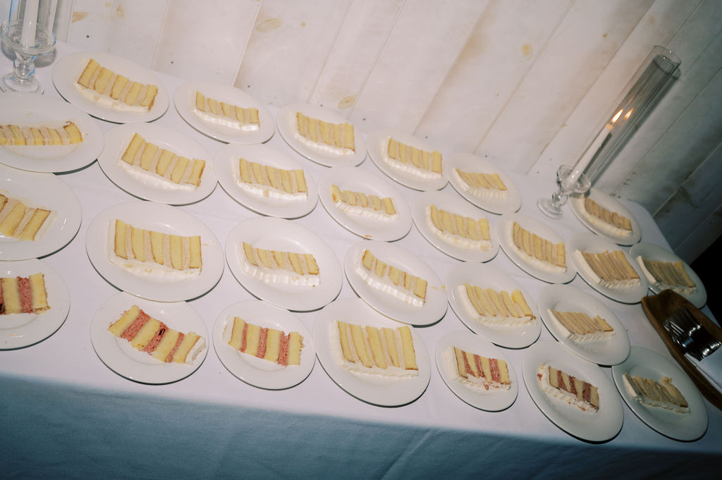 Several plates with slices of layered cake are arranged in rows on a white tablecloth next to a tall candleholder and a metal container.