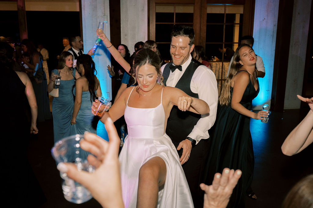 A bride in a white dress dances energetically with a man in formal wear at a wedding reception, surrounded by guests holding drinks.
