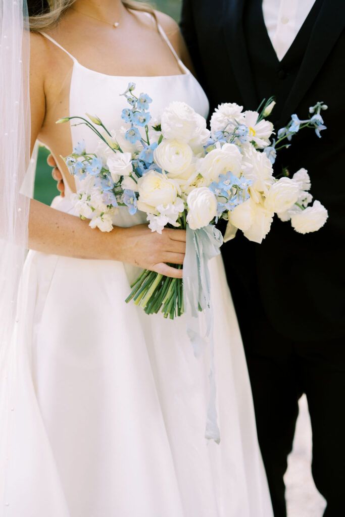 A bride in a white dress holds a bouquet of white and light blue flowers, standing next to a person in a black suit.