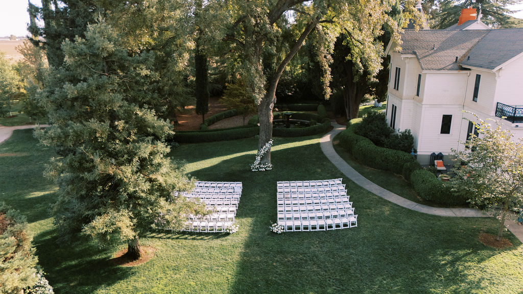 Aerial view of an outdoor wedding ceremony setup with rows of white chairs facing a tree, next to a large white house surrounded by greenery.